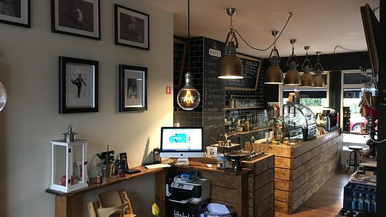 Ein gemütliches Café mit Holzmöbeln und einer Barista-Theke. An den Wänden hängen Bilder, und es gibt warme Beleuchtung von mehreren Lampen.