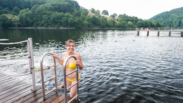 Eine Person in einem gelben Badeanzug steigt aus einem See auf einen Holzsteg. Im Hintergrund sind grüne Hügel und ein ruhiges Gewässer zu sehen.