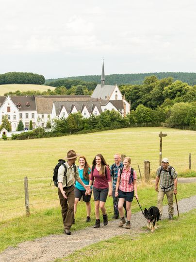 Eine Gruppe von Wanderern geht einen Kiesweg entlang, umgeben von grünen Wiesen. Im Hintergrund ist ein hübsches Gebäude mit einem Kirchturm zu sehen.