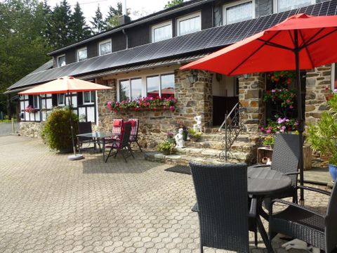 Ein gemütliches Restaurant mit einem Steinfassade, umgeben von bunten Blumen. Auf dem Platz stehen Tische mit roten Sonnenschirmen.