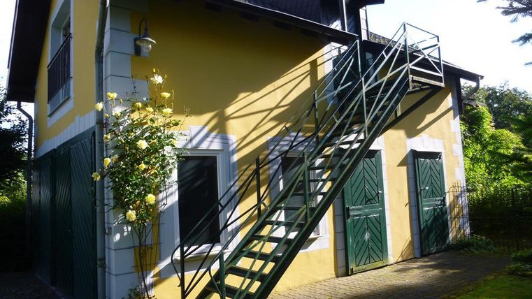 Ein charmantes Haus mit gelber Fassade und grünen Türen. Eine Treppe führt zur oberen Etage, umgeben von einer grünen Landschaft.