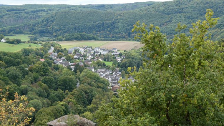 Eine malerische Landschaft mit Hügeln und einem kleinen Dorf im Tal. Im Vordergrund sind Bäume und Felsen sichtbar.
