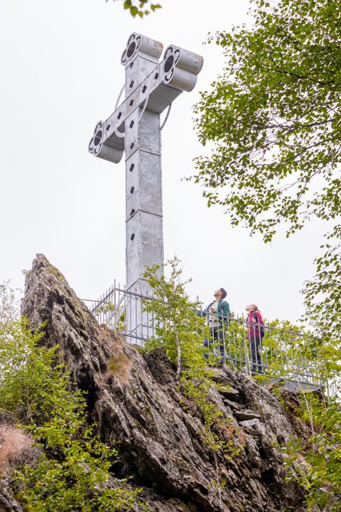 Ein großes, modernes Kreuz steht auf einem Felsen im Wald. Zwei Personen stehen nahe dem Kreuz und genießen die Aussicht.