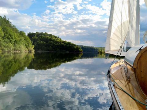 Ein Segelboot fährt auf einem ruhigen Gewässer, umgeben von grünen Hügeln. Der Himmel spiegelt sich klar im Wasser.