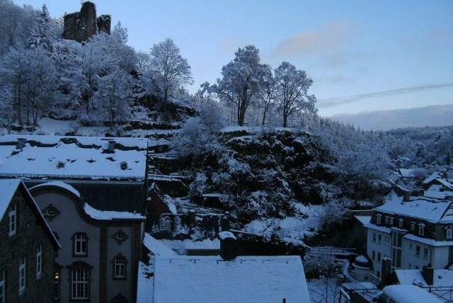 Eine winterliche Landschaft mit schneebedeckten Bäumen und einem Hügel im Hintergrund. Auf dem Hügel steht eine alte Burg, umgeben von ruhigen Häusern.