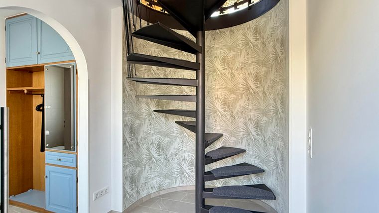 Eine moderne Wendeltreppe in einem hellen Raum. Im Hintergrund sind eine blaue Schrankwand und eine floral-musternde Wand sichtbar.