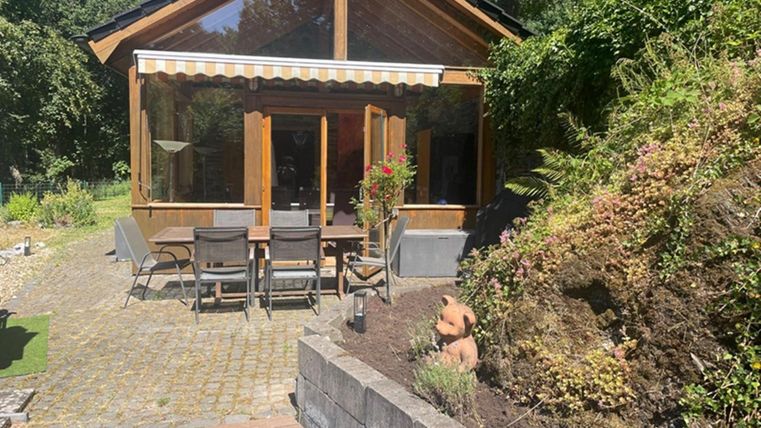 Ein gemütliches Gartenhaus mit großen Fenstern und einem sichtbaren Außenbereich. Es gibt einen Tisch mit Stühlen und eine gut gestaltete Landschaft rundherum.
