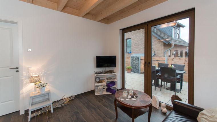 Ein modernes Wohnzimmer mit Holzbalkendecke und großen Fenstern. Es gibt einen Fernseher, einen Tisch und einen Zugang zu einem Außenbereich.