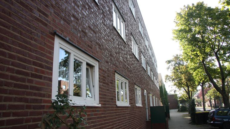 Ein mehrstöckiges Backsteingebäude mit Fenstern an der Seitenansicht. Im Vordergrund sind Bäume und ein Weg zu sehen.