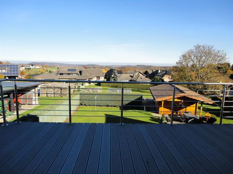Ein Blick von einer Terrasse auf eine ländliche Gegen, umgeben von Häusern und grünen Gärten. Der Himmel ist klar und blau, was für eine angenehme Atmosphäre sorgt.