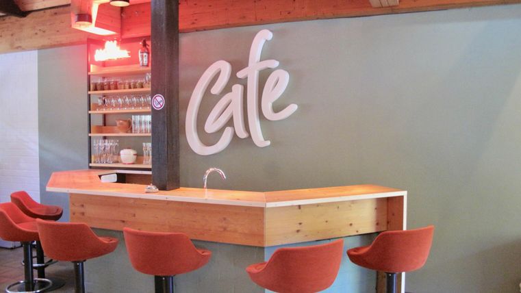 Ein moderner Cafébereich mit einer Holztheke und orangesfarbigen Stühlen. An der Wand steht das Wort "Café" in stilvollen Buchstaben.