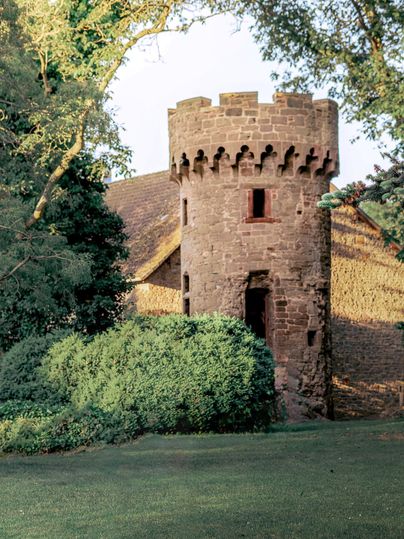 Ein kleiner historischer Turm aus Stein umgeben von grünen Büschen. Im Hintergrund ist ein altes Gebäude und Bäume zu sehen.