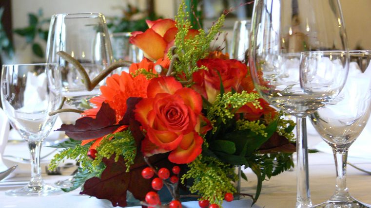 Ein schön gedeckter Tisch mit Weingläsern und einem floralen Arrangements aus orangefarbenen Rosen. Die Tischdekoration verleiht eine festliche Atmosphäre.
