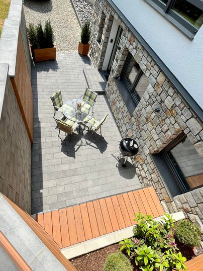 Eine moderne Terrasse mit einem Tisch und Stühlen. Im Hintergrund sieht man ein Steingebäude und einen Grill.