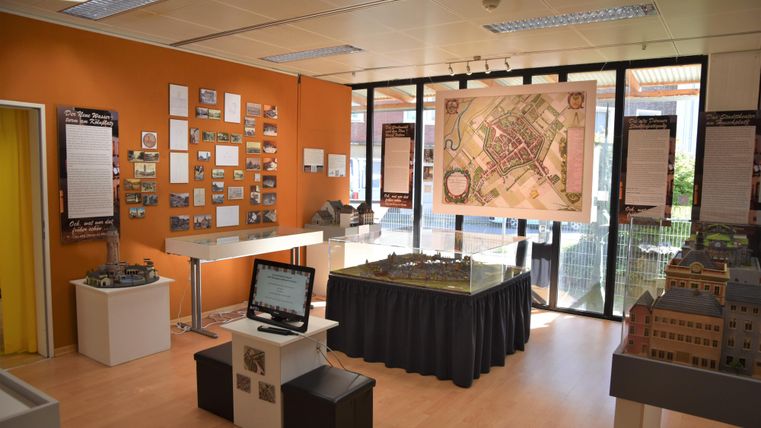 Ein moderner Ausstellungsraum mit orangefarbenen Wänden. Es gibt eine Karte, Modelle und Informationstafeln über die Geschichte.