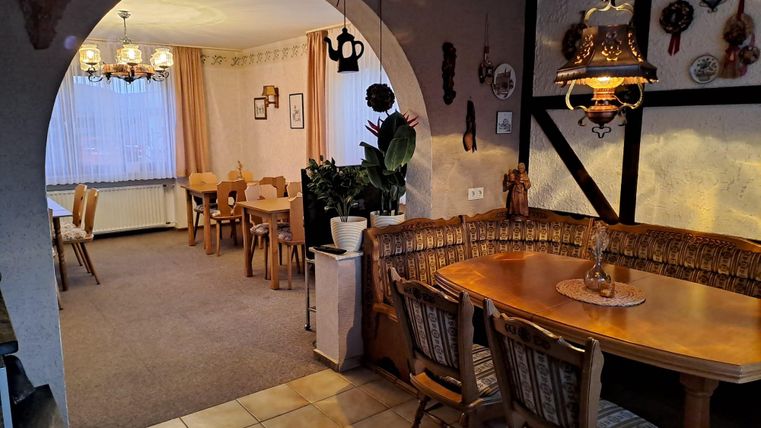 Ein gemütlicher Essbereich mit Holzstühlen und einem runden Tisch. Im Hintergrund sind weitere Sitzgelegenheiten und angenehmes Licht zu sehen.