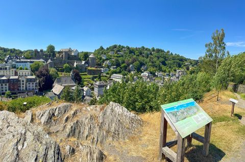 Eine Aussicht auf eine malerische Stadt mit grünen Hügeln im Hintergrund. Im Vordergrund steht ein Informationstafel auf einem Felsen.
