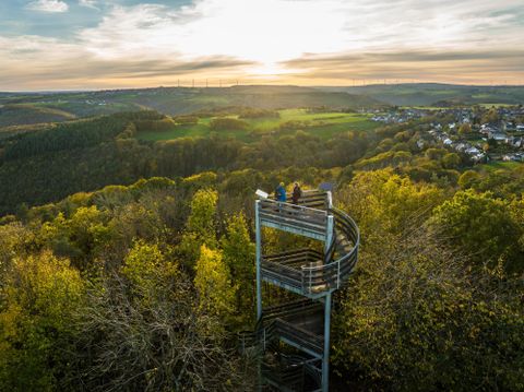 Eine Aussichtsplattform in der Natur mit Blick auf grüne Hügel und ein malerisches Dorf. Die Sonne geht hinter den Bergen unter.