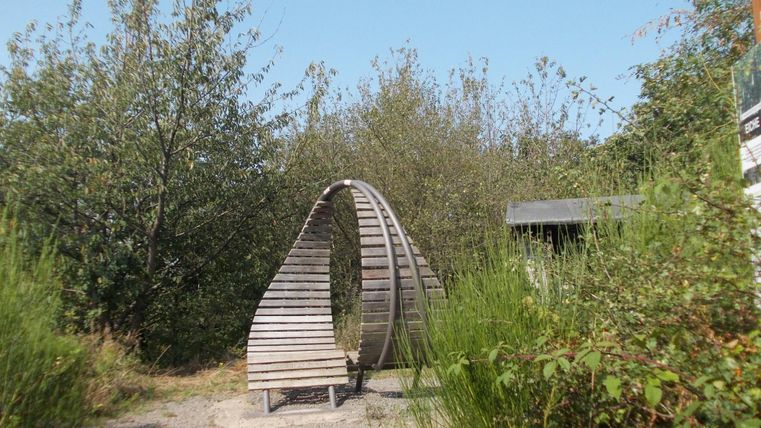 Eine Holzskulptur in Form eines Bogens steht in einer grünen Umgebung mit Sträuchern und Bäumen. Der Himmel ist blau und klar.
