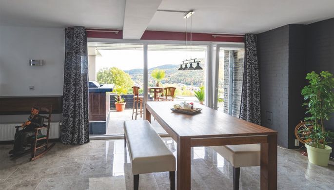 Ein modernes Esszimmer mit einem großen Holztisch und bequemen Bänken. Durch die großen Fenster blickt man auf eine grüne Landschaft.