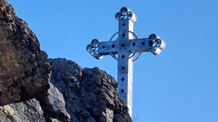 Ein großes, silbernes Kreuz steht auf einem Felsen unter einem klaren blauen Himmel. Die Umgebung ist grün und felsig.