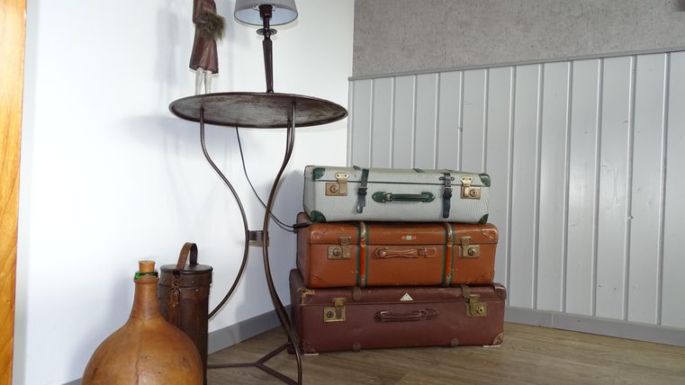 Ein vintage Tisch mit einer Lampe steht neben einem Stapel von alten Koffern. Darunter befinden sich eine große Flasche und ein kleiner Behälter.