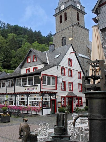 Eine malerische Dorfstraße mit traditionellen Fachwerkhäusern und einer Kirchturm im Hintergrund. Im Vordergrund steht ein Brunnen mit einer kindlichen Statue.