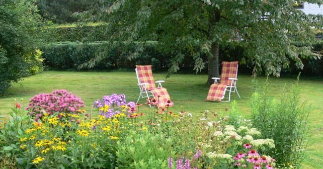 Ein schöner Garten mit bunten Blumen und zwei Liegestühlen unter einem Baum. Der Rasen ist grün und einladend.