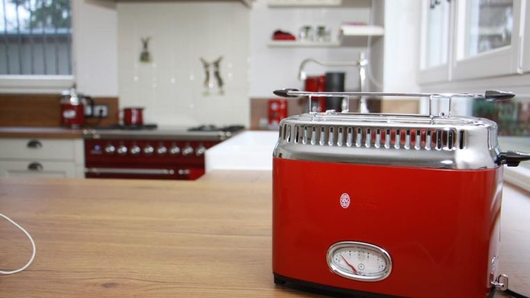 Ein roter Toaster steht auf einem Holzbrett in einer modernen Küche. Im Hintergrund sind ein Herd und Regale mit Geschirr sichtbar.