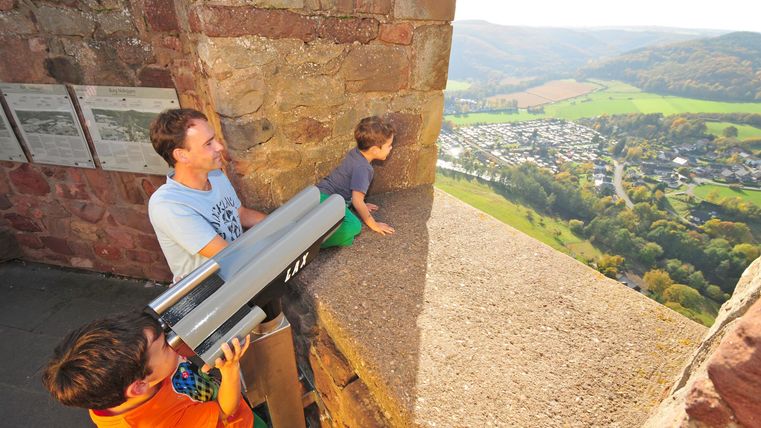 Ein Vater und zwei Kinder schauen durch ein Fernrohr von einer erhöhten Stelle. Im Hintergrund ist die schöne Landschaft mit einem Dorf und Hügeln zu sehen.