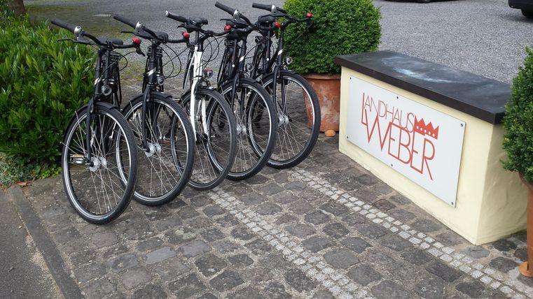Eine Gruppe von Fahrrädern steht vor einem Geschäftsschild mit dem Namen "Weber". Der Bereich ist von Pflanzen und einer Pflastersteinstraße umgeben.