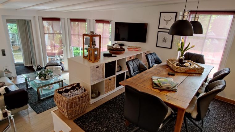 Ein gemütlicher Wohnraum mit einem Esstisch und einer modernen Couch. Große Fenster lassen viel Licht hereingelassen, und dekorative Elemente schaffen eine einladende Atmosphäre.