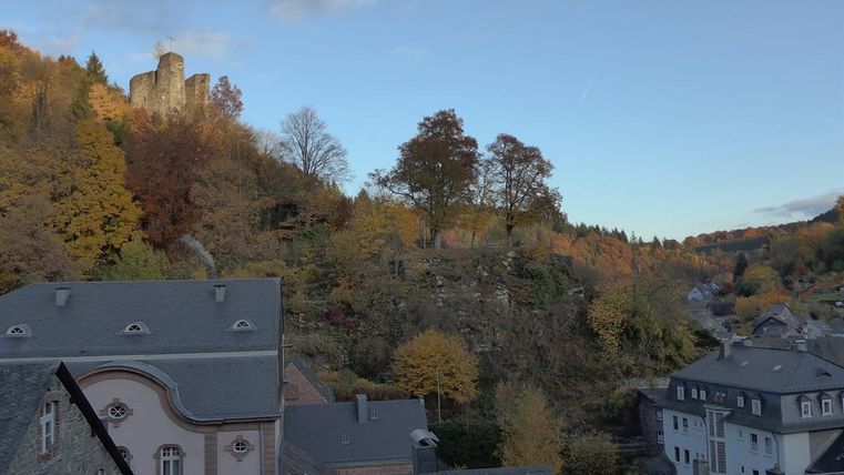 Eine malerische Landschaft mit buntem Herbstlaub und einer alten Burg auf einem Hügel. Im Vordergrund sind einige Häuser und Bäume zu sehen.