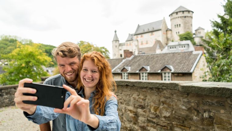 Ein Paar macht ein Selfie vor einer alten Burg. Im Hintergrund sind Bäume und historische Gebäude zu sehen.