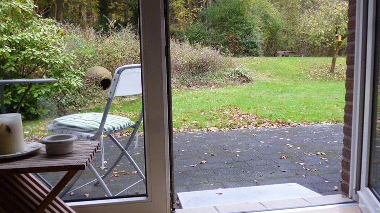 Ein Blick aus einem Fenster auf einen Garten mit Rasen und Sträuchern. Im Vordergrund steht ein Stuhl und ein Tisch.