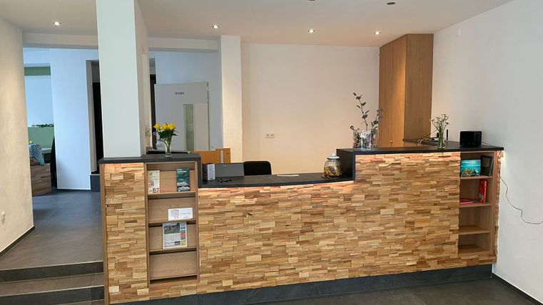 Ein moderner Empfangsbereich mit einer Holztheke und einem hellen, einladenden Raum. Die Wände sind in neutralen Farben gehalten, und es gibt einige Pflanzen als Dekoration.