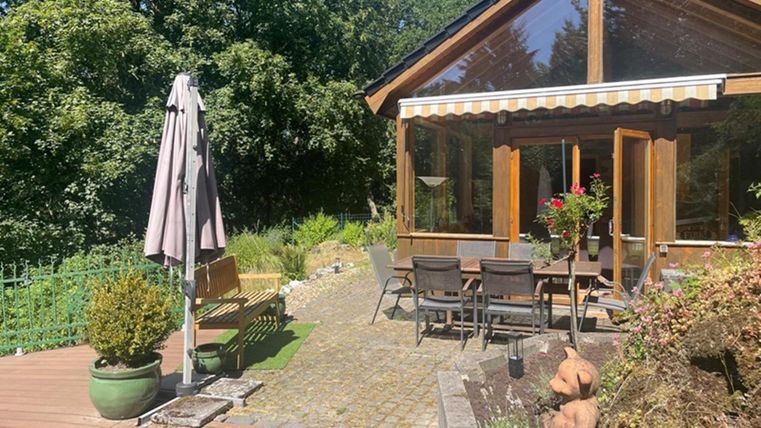 Ein gemütliches Holzhaus mit großen Fenstern und einer Terrasse. Der Außenbereich ist von Pflanzen und einem Sonnenschirm umgeben.