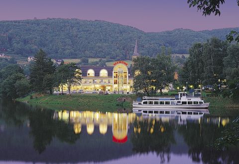 Ein malerischer Blick auf ein Gebäude am Fluss mit sanften Hügeln im Hintergrund. Auf dem Wasser spiegelt sich das Licht des Gebäudes.