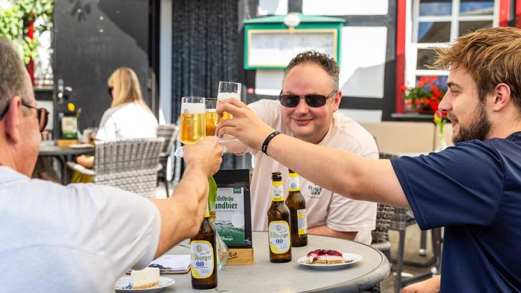 Eine fröhliche Runde von Freunden, die auf ein Bier anstoßen. Im Hintergrund sind Stühle und Tische in einem einladenden Biergarten zu sehen.