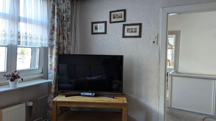 Ein Wohnzimmer mit einem Fernseher auf einem Holzständer. An der Wand hängen einige Bilder und ein Fenster mit Vorhängen ist ebenfalls sichtbar.