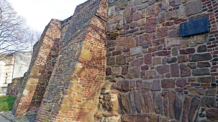 Eine alte Mauer aus Stein mit einer unregelmäßigen Struktur. Die Mauer zeigt Spuren der Geschichte und ist von Bäumen umgeben.