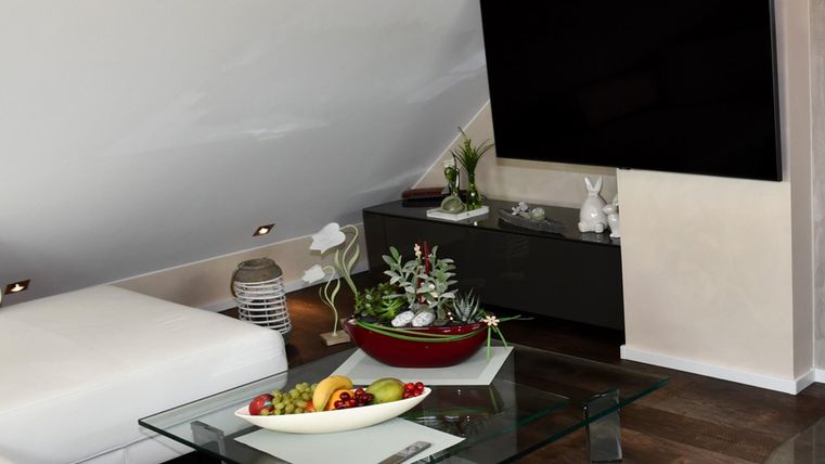 Ein modernes Wohnzimmer mit einem großen Fernseher und einem Glastisch. Auf dem Tisch befindet sich eine Obstschale und einige Pflanzen.