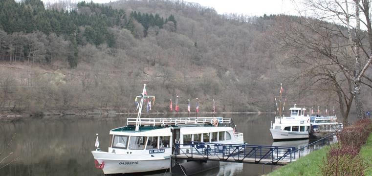 Zwei Ausflugsschiffe liegen an einem Steg am Fluss. Im Hintergrund sind bewaldete Hügel zu sehen.