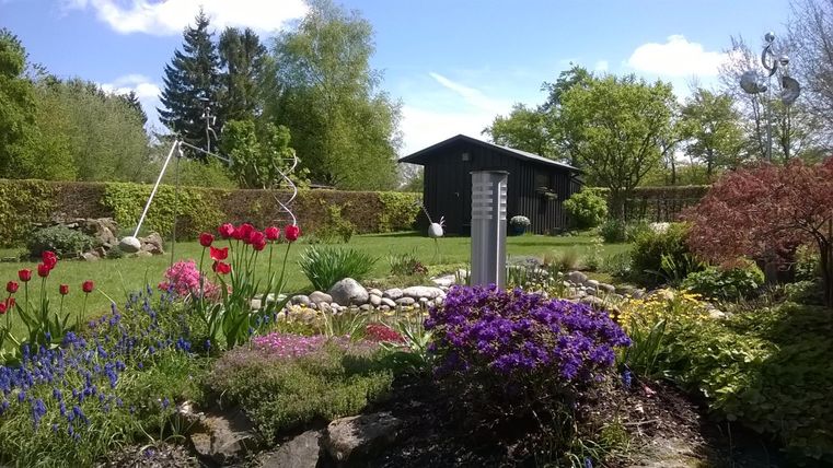 Ein schöner Garten mit bunten Blumen und einem Holzhaus im Hintergrund. Die Sonne scheint und die Bäume umgeben die Szene.