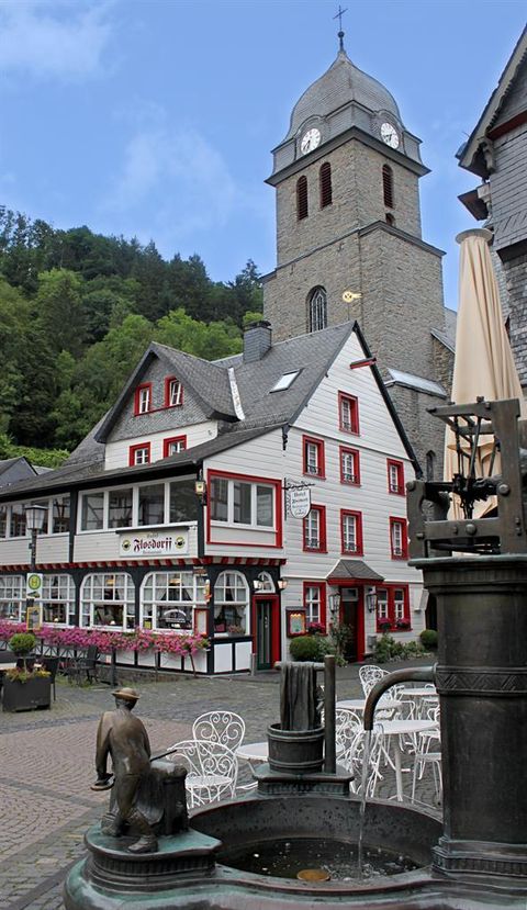 Eine malerische Dorfstraße mit traditionellen Fachwerkhäusern und einer Kirchturm im Hintergrund. Im Vordergrund steht ein Brunnen mit einer kindlichen Statue.