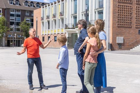 Eine Gruppe von fünf Personen steht in einem öffentlichen Raum. Sie scheinen miteinander zu sprechen und zu lachen, während im Hintergrund moderne Gebäude sichtbar sind.