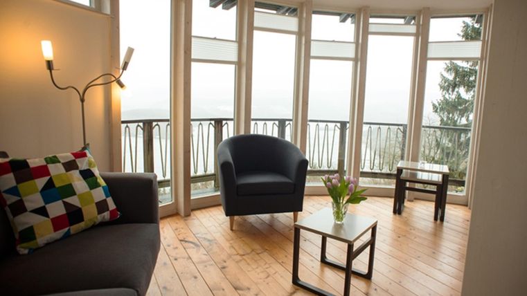 Ein gemütlicher Raum mit großen Fenstern und einem Blick auf die Landschaft. Es gibt eine Sofaecke mit bunten Kissen und einen kleinen Tisch mit frischen Blumen.