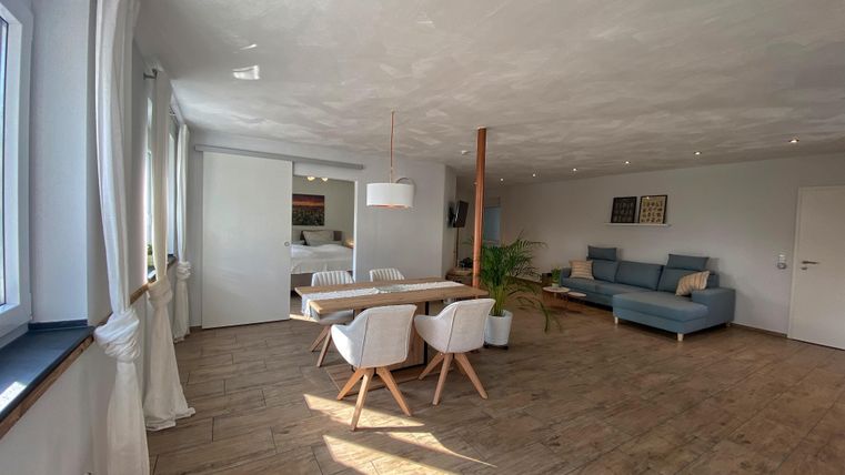 Ein helles und modernes Wohn- und Esszimmer mit einem großen Tisch und weißen Stühlen. Der Raum hat eine gemütliche Atmosphäre mit einer blauen Couch und großen Fenstern.