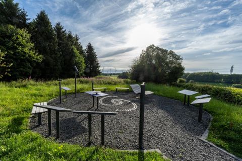 Ein runder Bereich mit Tischen und Schautafeln in einer grünen Landschaft. Im Hintergrund sind Bäume und ein klarer Himmel sichtbar.