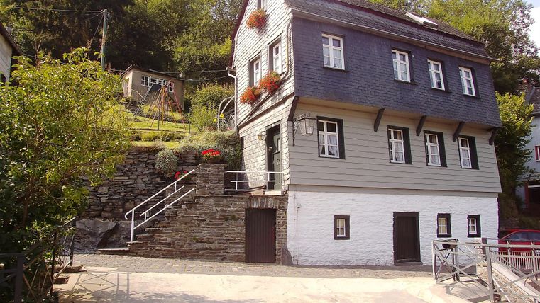 Ein historisches Haus mit grauer Fassade und weißen Akzenten. Es steht in einer grün bewachsenen Umgebung mit Stufen und einem kleinen Garten.